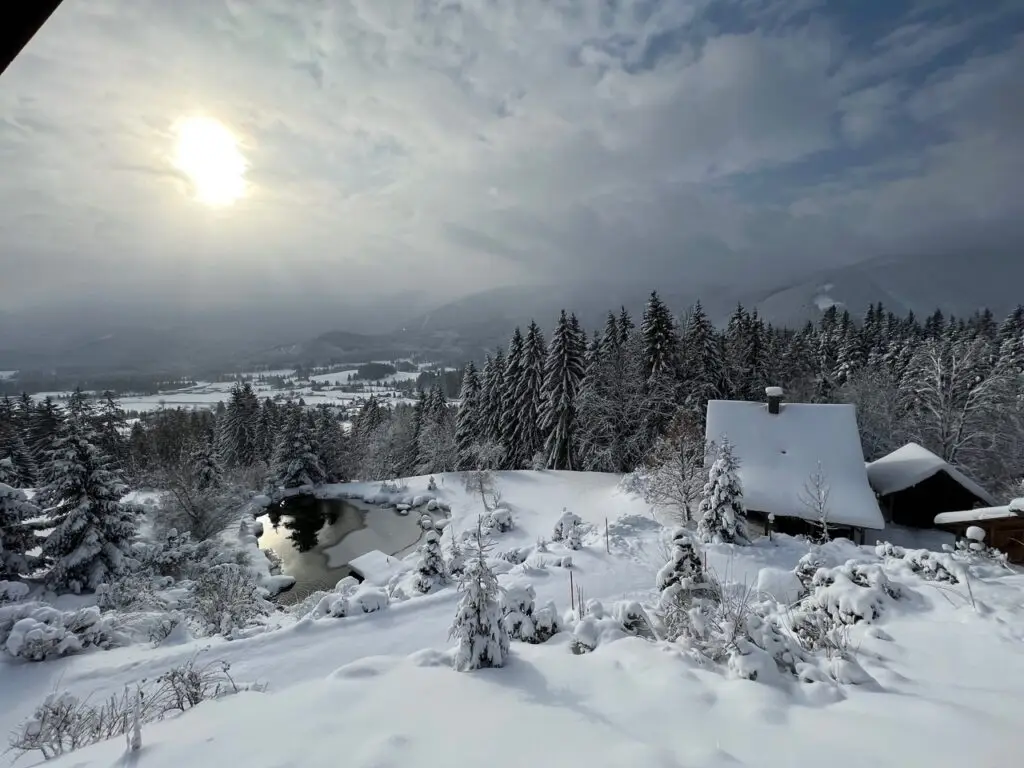 Eine schneebedeckte Landschaft mit einer Hütte an einem Teich, umgeben von Pinienbäumen. Weiches Licht taucht die winterliche Szenerie und die fernen Berge in ein Licht, das an die ruhige Atmosphäre der Lichtquellalm, der Heimat von Dr. Töths Licht-Quanten Forschung, erinnert.