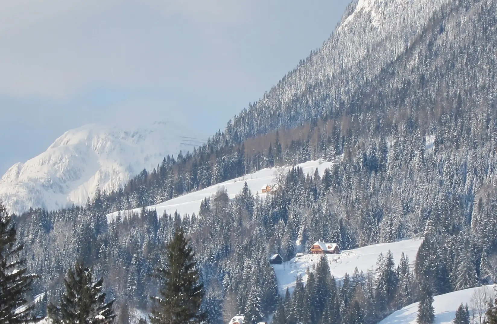 Schneebedeckte Berge mit dichten, immergrünen Wäldern umgeben verstreute Hütten unter einem klaren, blauen Himmel. Diese ruhige, winterliche Landschaft ist ein echter Kraftort in der Steiermark, der vom Sonnenlicht erhellt wird und zur friedlichen Besinnung inmitten der fernen Gipfel einlädt.