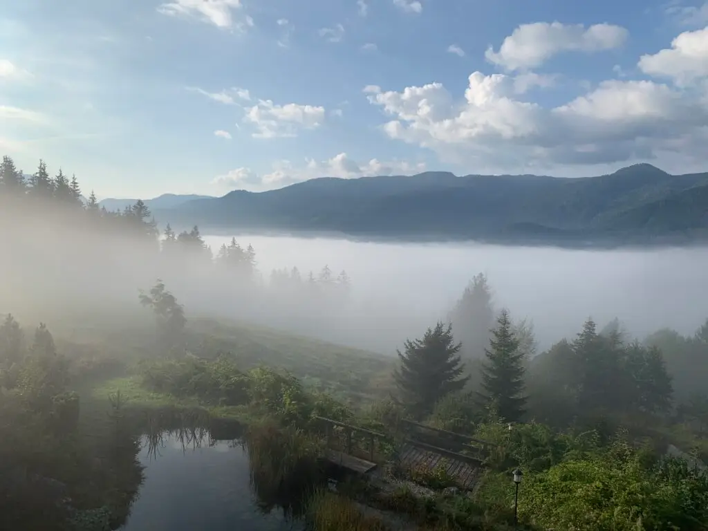 Eine neblige Landschaft mit dichtem Nebel in einem Tal in der Nähe der Licht-Quell-Alm, über die Hügel verstreute Kiefern, ein kleiner Teich, eine Holzbrücke und Berge unter einem teilweise bewölkten blauen Himmel - ein wahrer Spiritueller Rückzugsort Alpen.