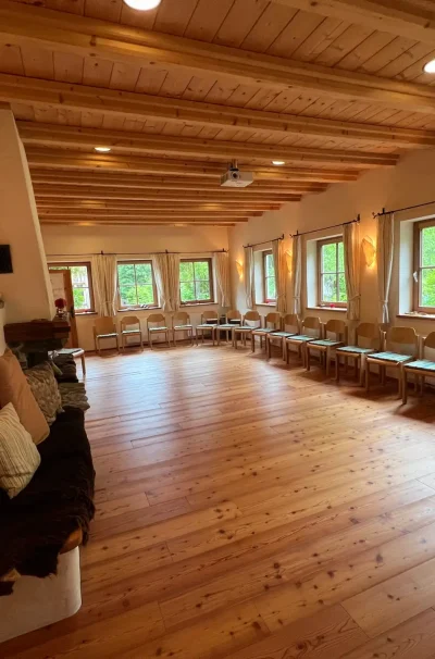Ein geräumiger, gemütlicher Raum mit Holzböden und Deckenbalken, großen Fenstern und gepolsterten Sitzgelegenheiten - perfekt für ein Burnout Prävention Retreat oder als ruhiger Yoga Rückzugsort Alpen. Warmes natürliches Licht erfüllt den Raum und lädt zu Entspannung und Erneuerung ein.