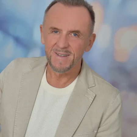 Ein lächelnder Mann mittleren Alters mit kurzen Haaren und gestutztem Bart, der einen hellbeigen Blazer über einem weißen Pullover trägt, posiert vor einem sanft verschwommenen blauen und lila Hintergrund, der seine Leidenschaft für die Naturheilkunde Salzkammergut und Blütenessenzen widerspiegelt.