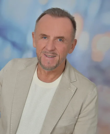 Ein lächelnder Mann mittleren Alters mit kurzen Haaren und gestutztem Bart, der einen hellbeigen Blazer über einem weißen Pullover trägt, posiert vor einem sanft verschwommenen blauen und lila Hintergrund, der seine Leidenschaft für die Naturheilkunde Salzkammergut und Blütenessenzen widerspiegelt.