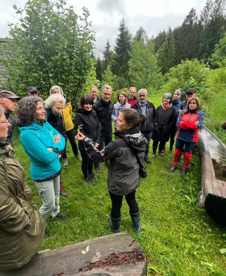 Eine Gruppe von Menschen steht im Freien in einem üppigen, grünen Bereich der Lichtquellalm und hört einer Frau zu, die eine Pflanze hält und über Blütenessenzen spricht. Sie sind in Jacken und Outdoor-Kleidung gekleidet, in der Nähe stehen Bäume und ein rustikaler Holztrog.