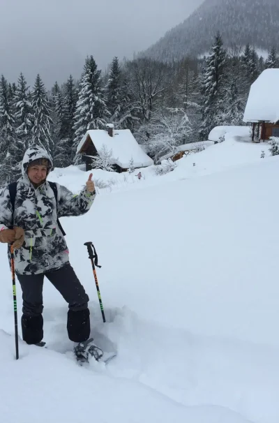 Eine Person in Winterausrüstung steht mit Skistöcken im tiefen Schnee in der Nähe einer einsamen Berghütte in der Steiermark, umgeben von Kiefern und Bergen unter einem bewölkten Himmel.