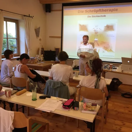 Ein Mann steht vorne in einem Klassenzimmer und hält einen Vortrag über "Die Schröpftherapie" im Rahmen einer Ganzheitsmedizin-Ausbildung. Auf den Tischen sind medizinische Hilfsmittel und Bücher zu sehen, und die Erwachsenen hören aufmerksam zu.