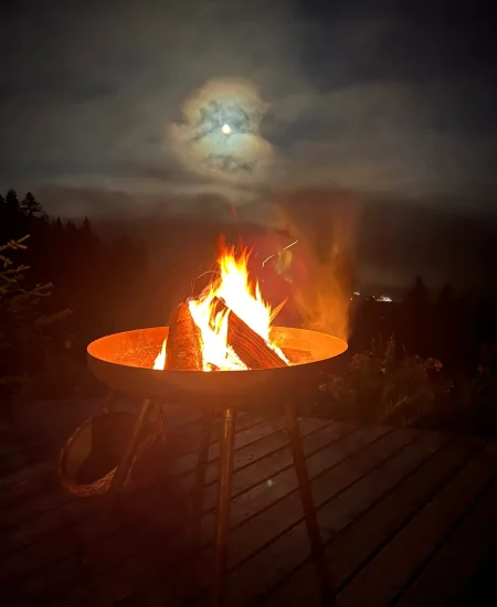 Eine Feuerstelle mit brennenden Holzscheiten auf einer Holzterrasse während eines nächtlichen Lichtquellalm-Seminars, mit einem leuchtenden Vollmond, der teilweise von Wolken und Bäumen im Hintergrund verdeckt wird, evoziert eine Atmosphäre von heiterer Licht-Quanten Forschung.