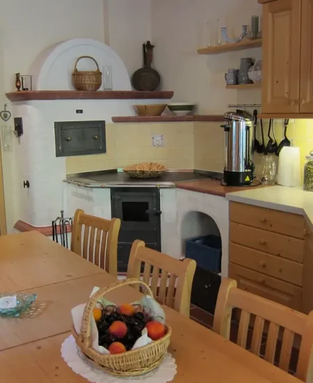 Eine gemütliche, rustikale Küche mit Holzschränken und -möbeln, einem langen Esstisch mit Obstkorb, Blütenessenzen von Dr. Töth in den Regalen, einem Herd, Arbeitsplattengeräten und einer sanften gelben Beleuchtung, die an die Lichtquellalm erinnert.