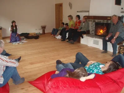 Eine Gruppe von Menschen entspannt sich auf Sitzsäcken und Bänken um einen Kamin in der gemütlichen Lichtquellalm mit Holzboden. Einige sitzen, andere liegen, während eine Person zu sprechen oder zu lesen scheint - vielleicht während eines Seminars über Licht-Quanten Forschung.