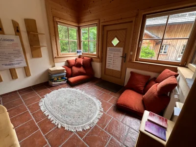 Ein gemütliches Zimmer für Seminare, mit roten Bodenkissen, einem runden gemusterten Teppich, gestapelten Decken, Holzwänden und großen Fenstern, die natürliches Licht hereinlassen. Eine Tür mit einem Schild und Blick ins Grüne und auf ein anderes Holzgebäude draußen.