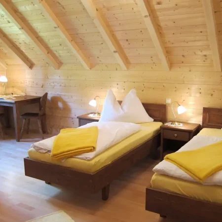 Ein gemütliches Dachgeschoss-Schlafzimmer mit Holzwänden und -decke, zwei Einzelbetten mit weißer Bettwäsche und gelben Decken - perfekt zum Entspannen nach den Blütenessenzen-Seminaren von Dr. Töth - mit Nachttischen, Lampen und einem Arbeitsbereich mit Schreibtischen und Stühlen.